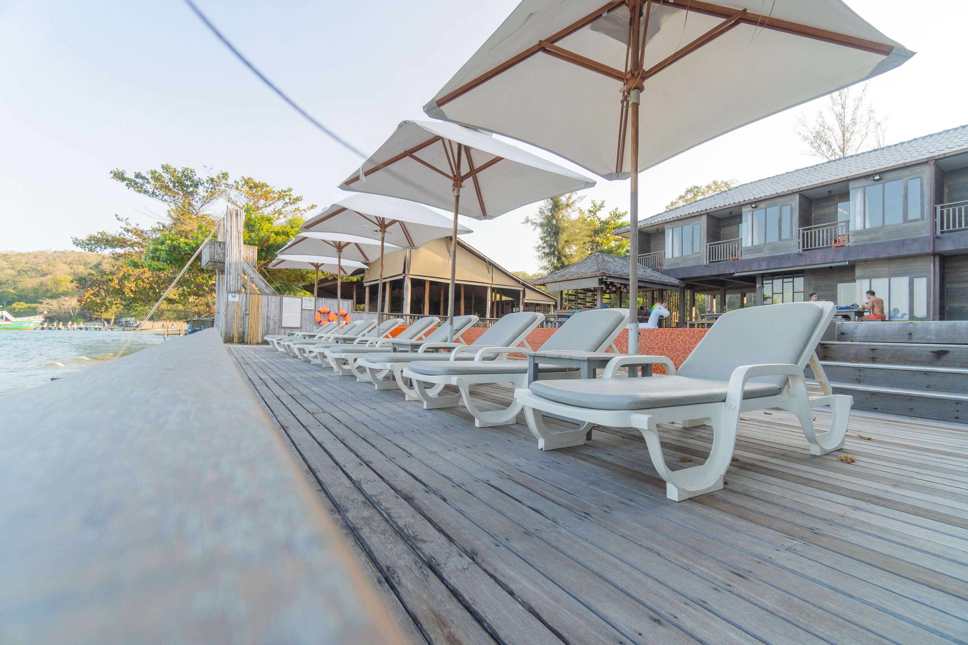 A wooden deck with white lounge chairs and umbrellas, overlooking a beach and a building with multiple balconies.