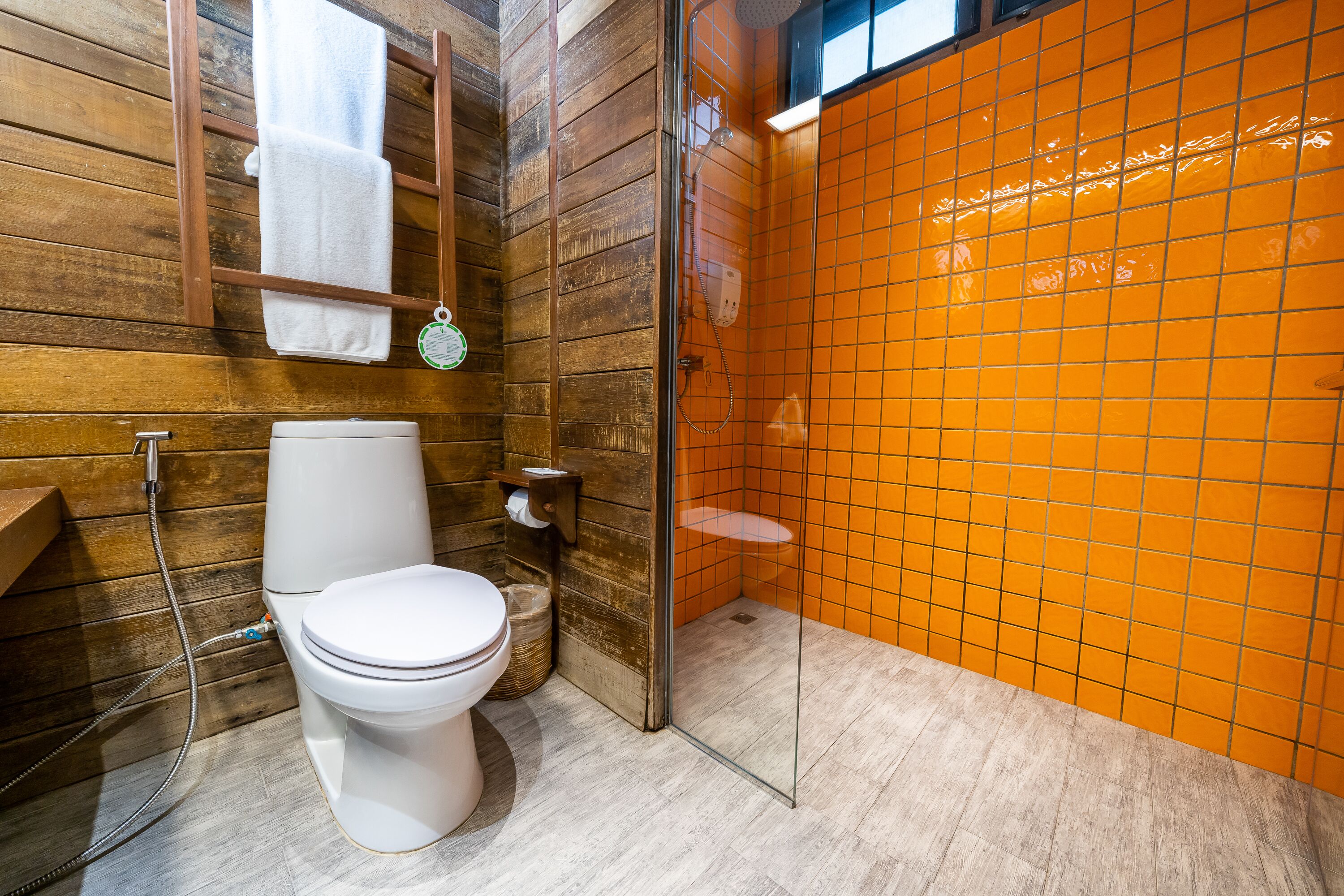 Et badeværelse med trægulv, et hvidt toilet, en orange flisevæg ved brusenichen og et glasbruseområde.