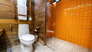 Et badeværelse med trægulv, et hvidt toilet, en orange flisevæg ved brusenichen og et glasbruseområde.