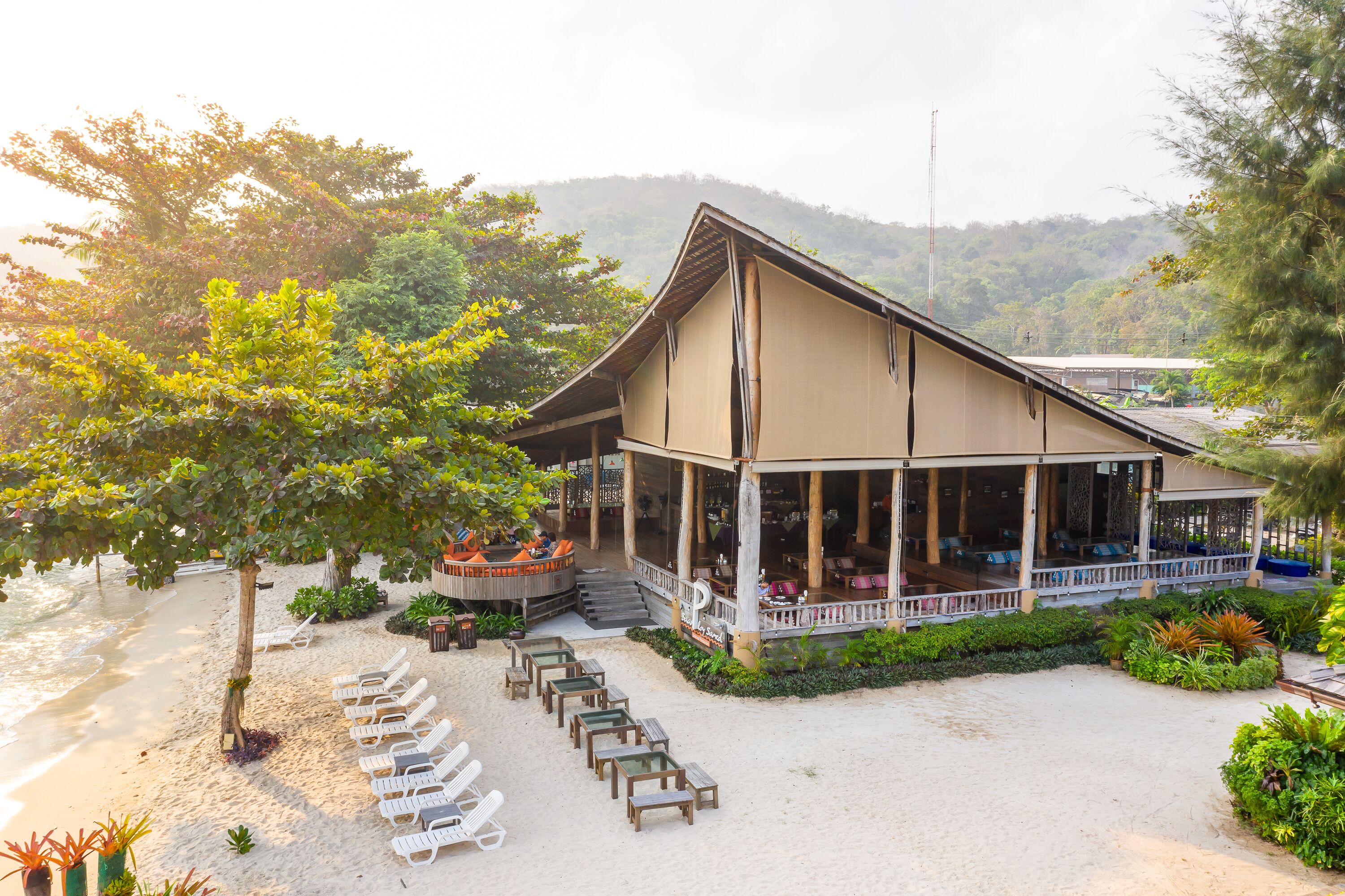 A beachside resort with a covered outdoor seating area, lounge chairs, and a view of the beach and hills.