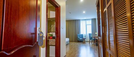 Un pasillo de hotel con una puerta de madera, un baño con ducha, una zona de estar con una silla y una mesa, y una ventana con cortinas.