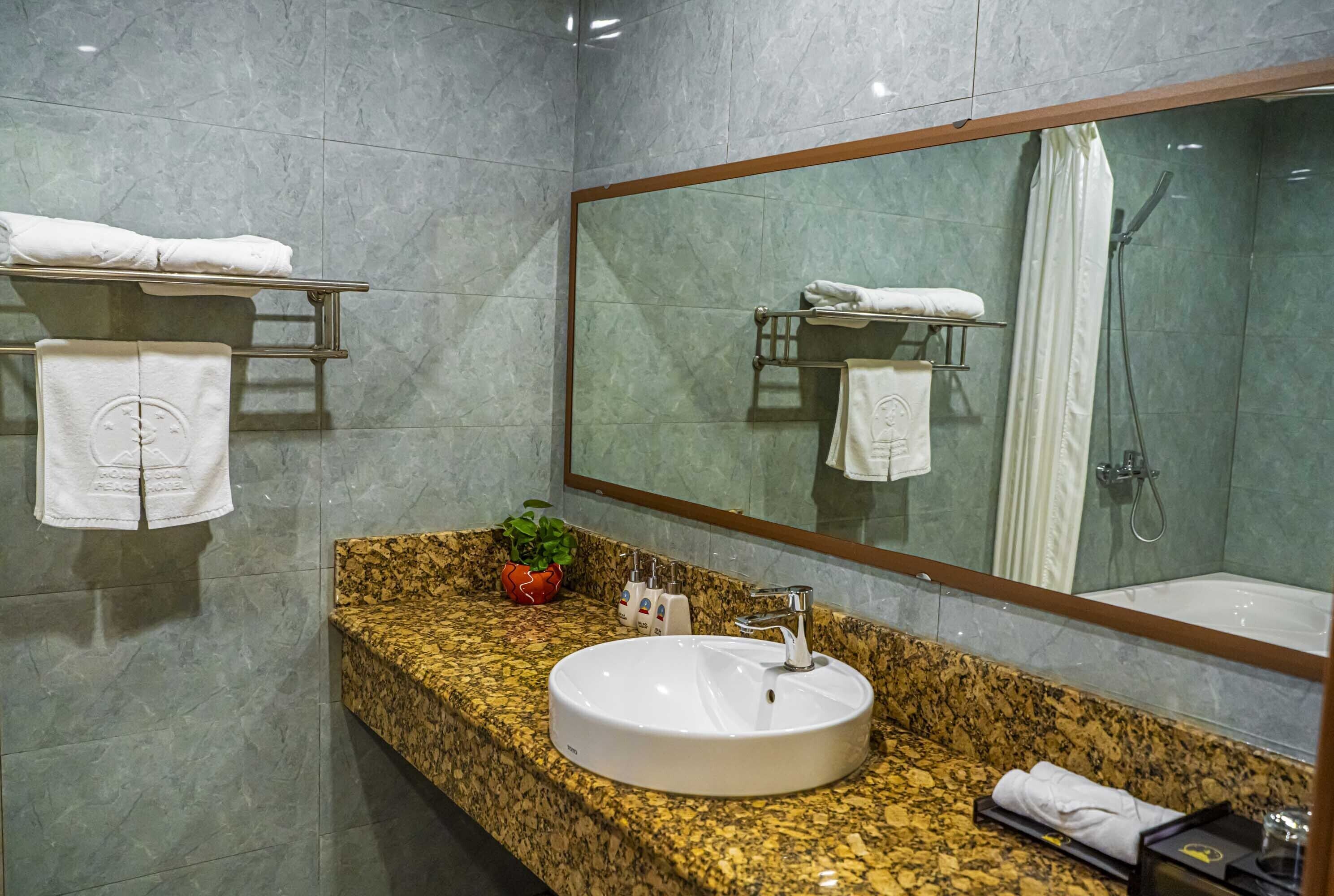 A modern bathroom with a granite countertop, a white sink, and a large mirror. There are towels on a shelf and a shower visible in the mirror.
