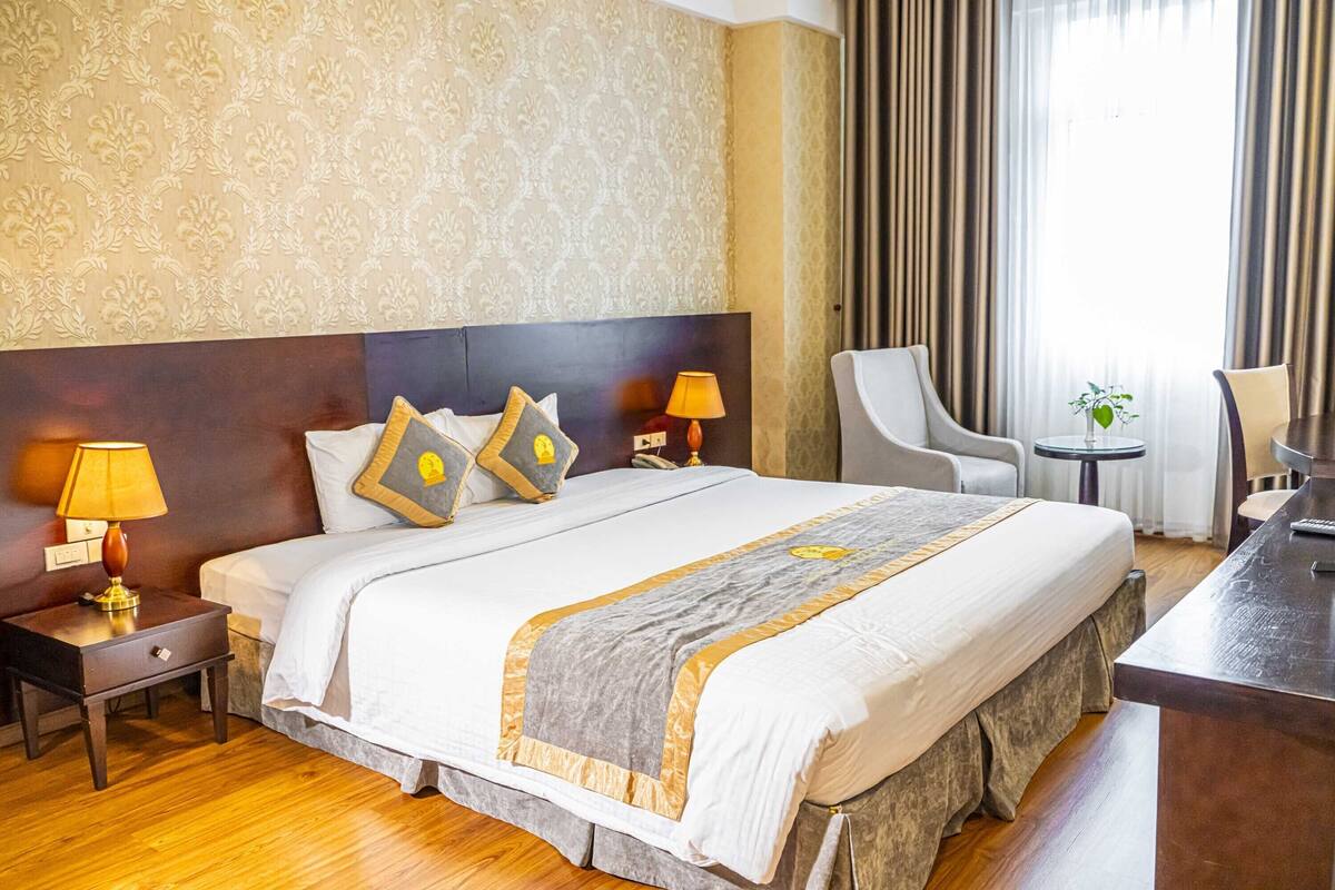 Ein Hotelzimmer mit einem großen Bett, zwei Nachttischen mit Lampen, einem Sessel, einem kleinen Tisch mit Pflanze und einem Schreibtisch mit Lampe.