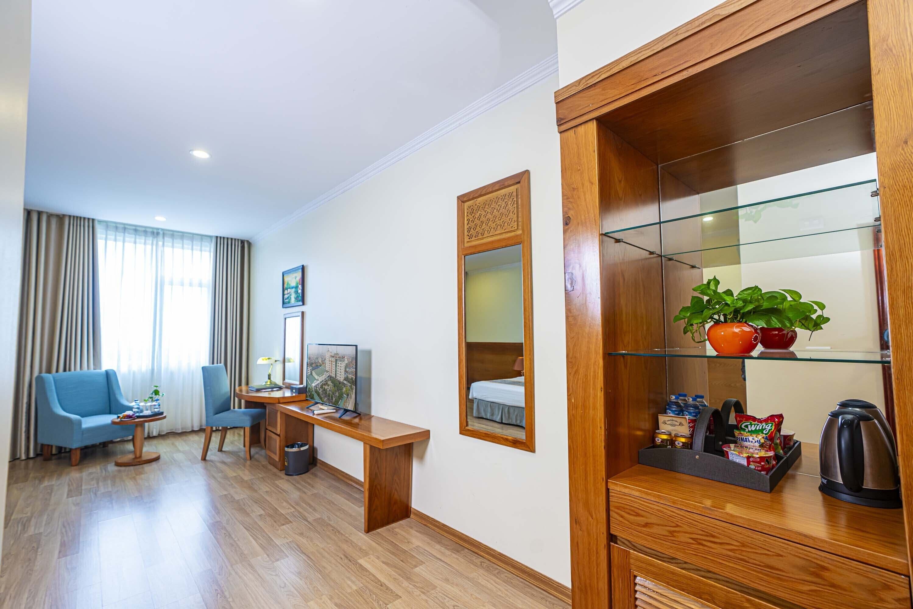 Habitación de hotel con escritorio de madera, sofá azul, pequeña mesa, espejo, estante con planta y cama con edredón blanco.