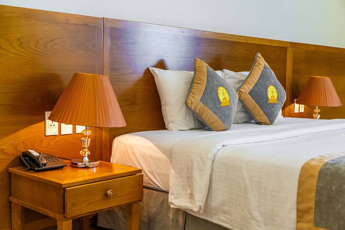 Ein Hotelzimmer mit Holz Kopfteil, zwei Nachttischlampen, einem Telefon auf einem Holznachttisch und einem Bett mit weißer Bettwäsche und Zierkissen.