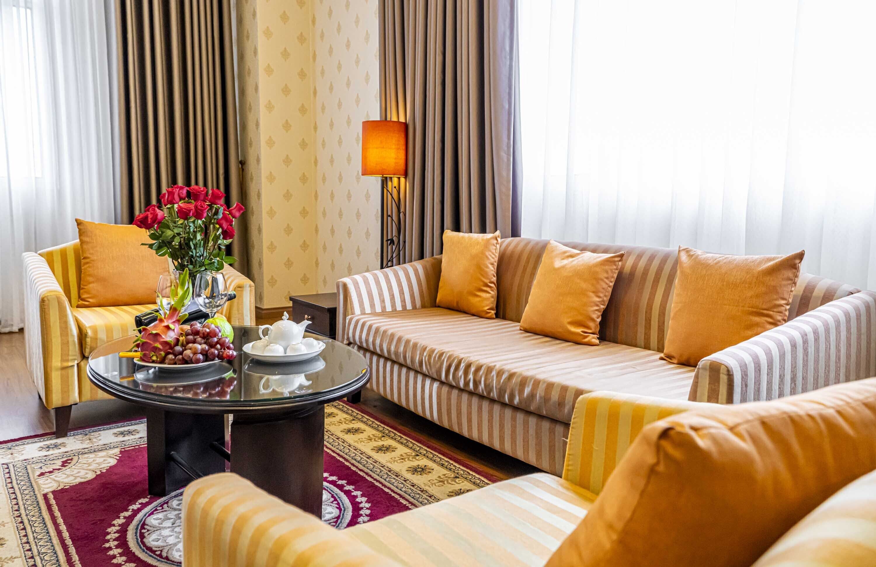 Una habitación de hotel con un sofá a rayas, un sillón amarillo, una mesa de centro redonda con un jarrón de rosas rojas y una lámpara.