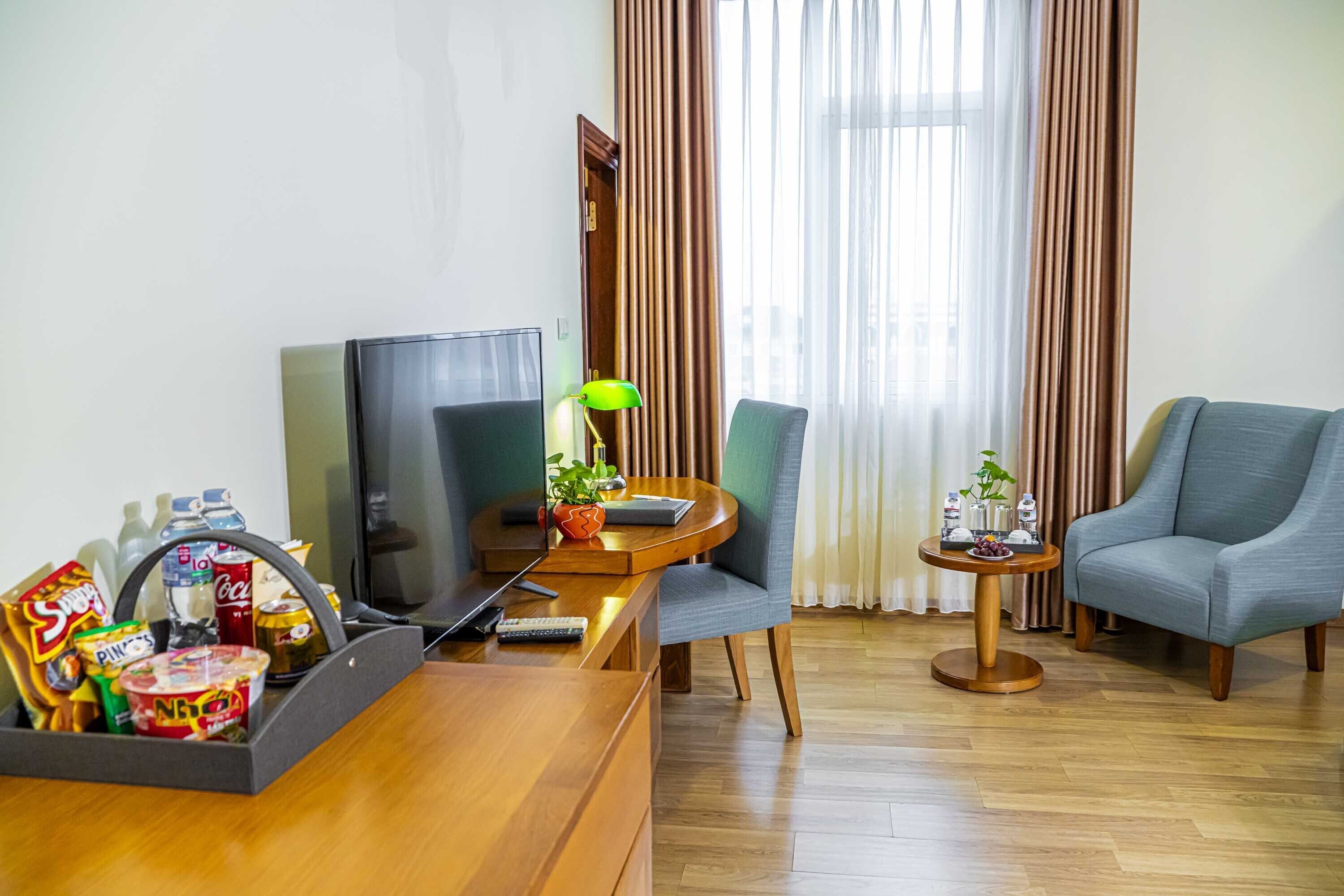 Une chambre d’hôtel avec un bureau en bois, une télévision, une chaise, une table d’appoint avec une lampe, et une vue sur l’extérieur à travers des rideaux.