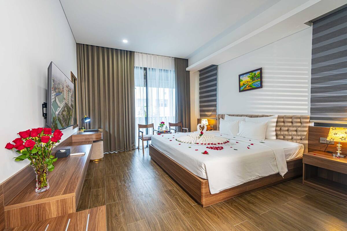 Ein modernes Hotelzimmer mit einem großen Bett, einem Schreibtisch mit Lampe, einem Sessel und einer Vase mit roten Rosen.