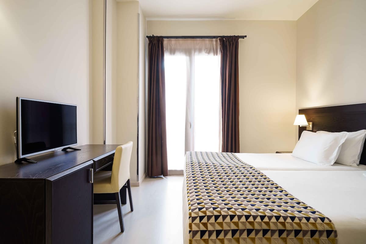 A hotel room with a bed, a desk with a TV, a chair, a lamp, and a window with curtains.