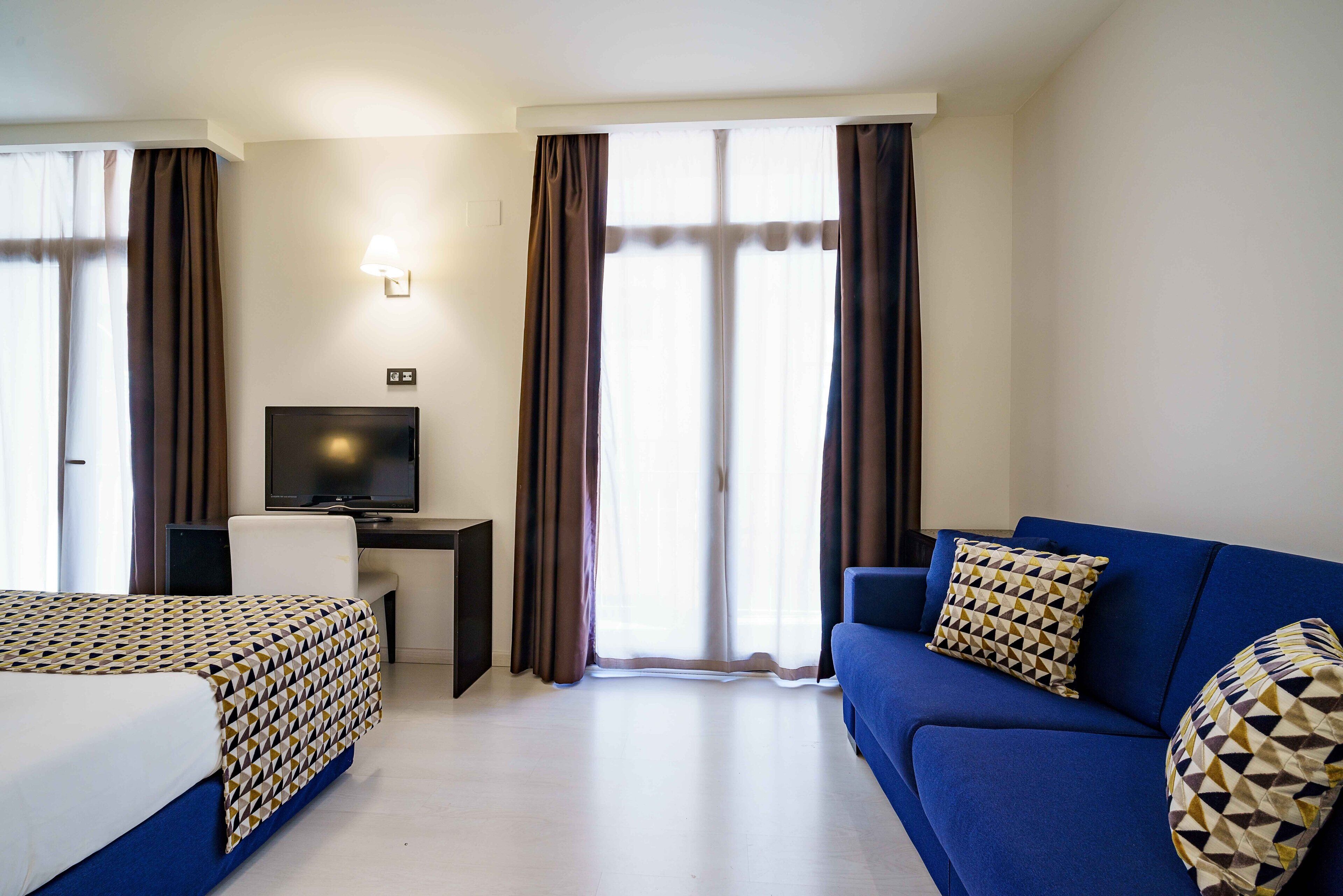 A hotel room with a bed, a desk with a television, a blue sofa, and curtains.
