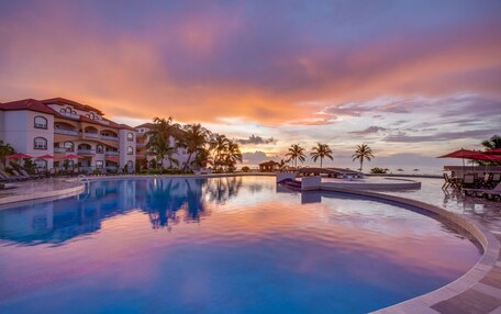 A resort pool with a sunset view, palm trees, and a building with balconies.