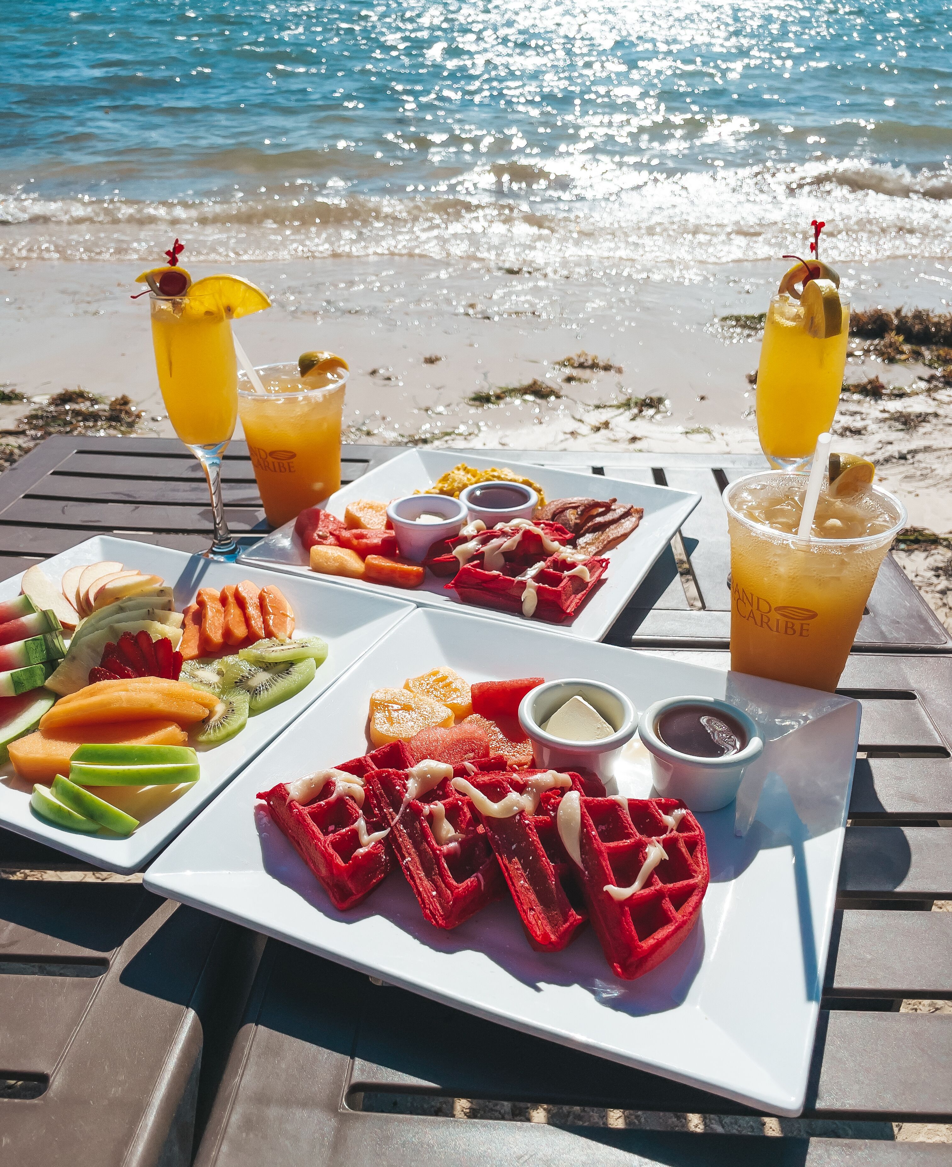 A beachside table with a variety of fruits, waffles, and drinks, set against a backdrop of the ocean.