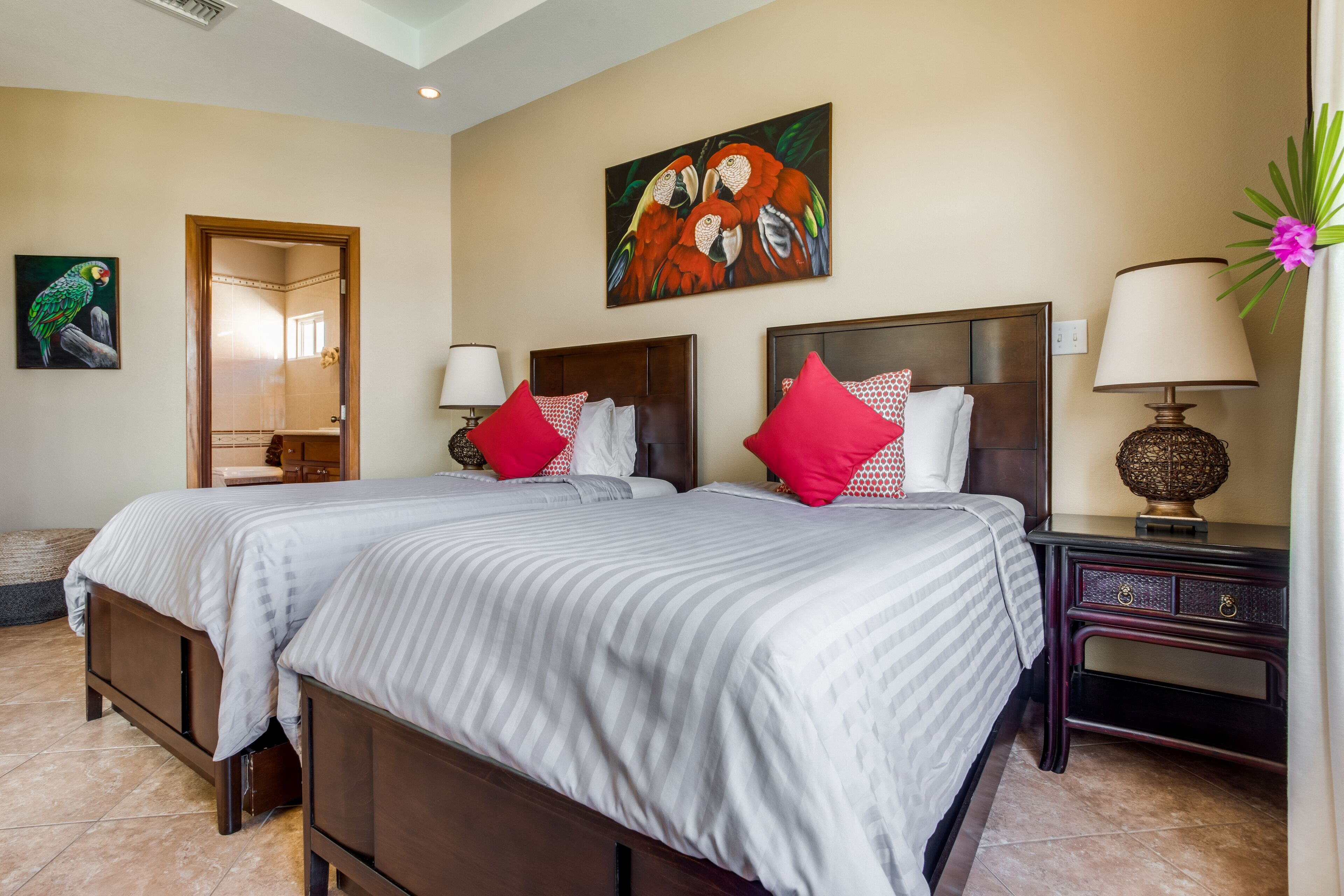 A hotel room with two beds, a wooden headboard, a nightstand, a lamp, and a painting of parrots on the wall.