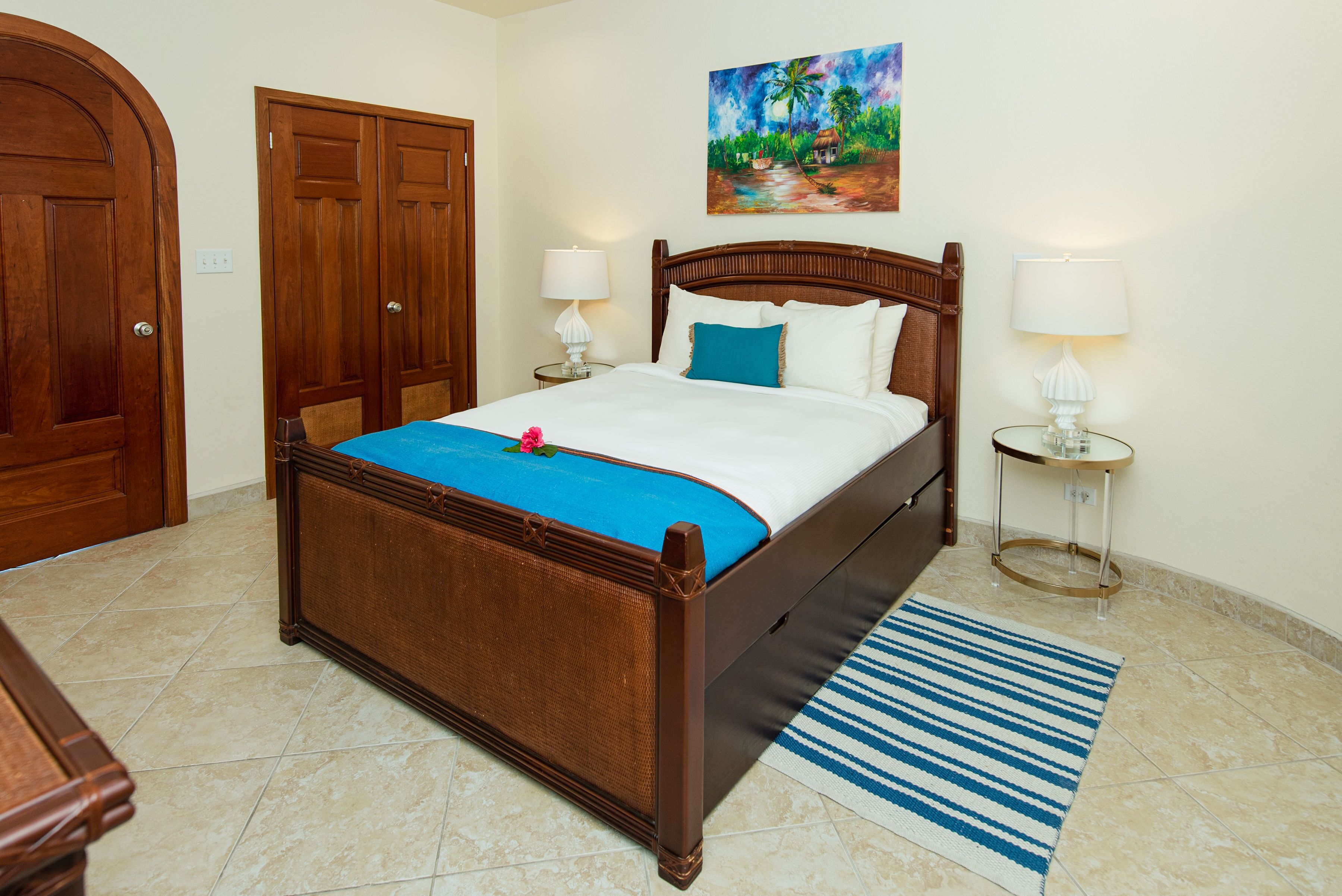A hotel room with a wooden bed, a blue striped rug, and a painting of a tropical scene.