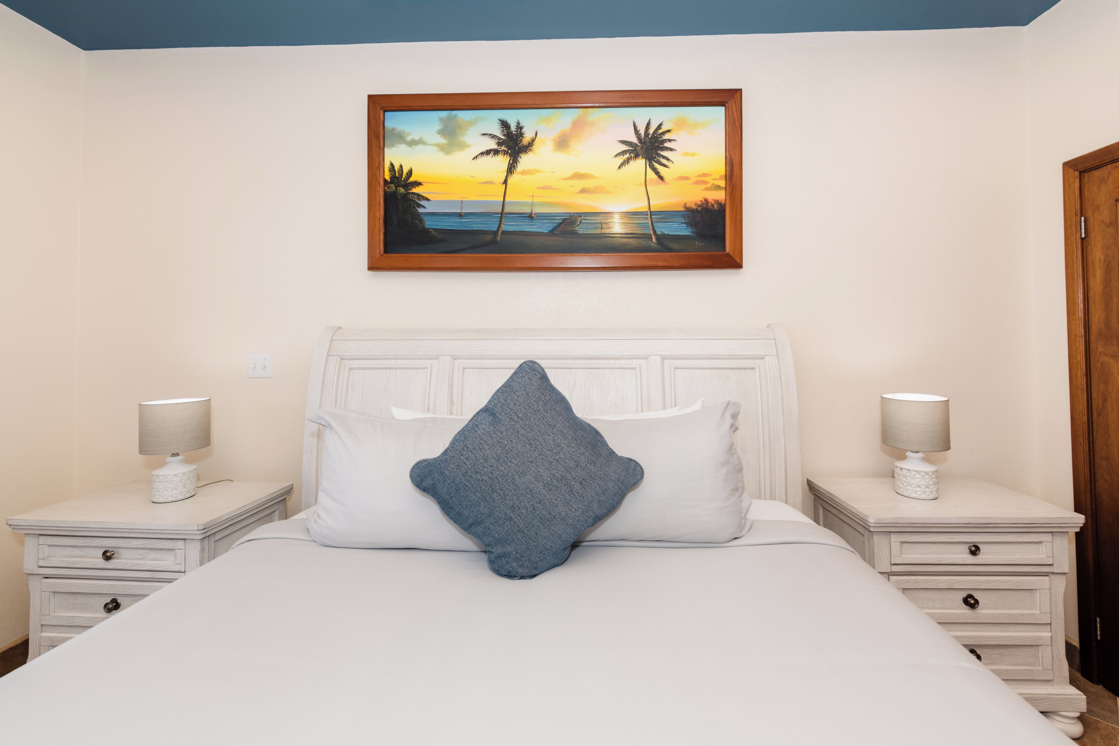 A neatly made bed with a blue pillow, two bedside tables with lamps, and a framed painting of a sunset over the ocean.