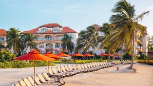 A beachside hotel with rows of sun loungers and red umbrellas, palm trees, and a building with a red roof. - Grand Caribe Belize (San Pedro)