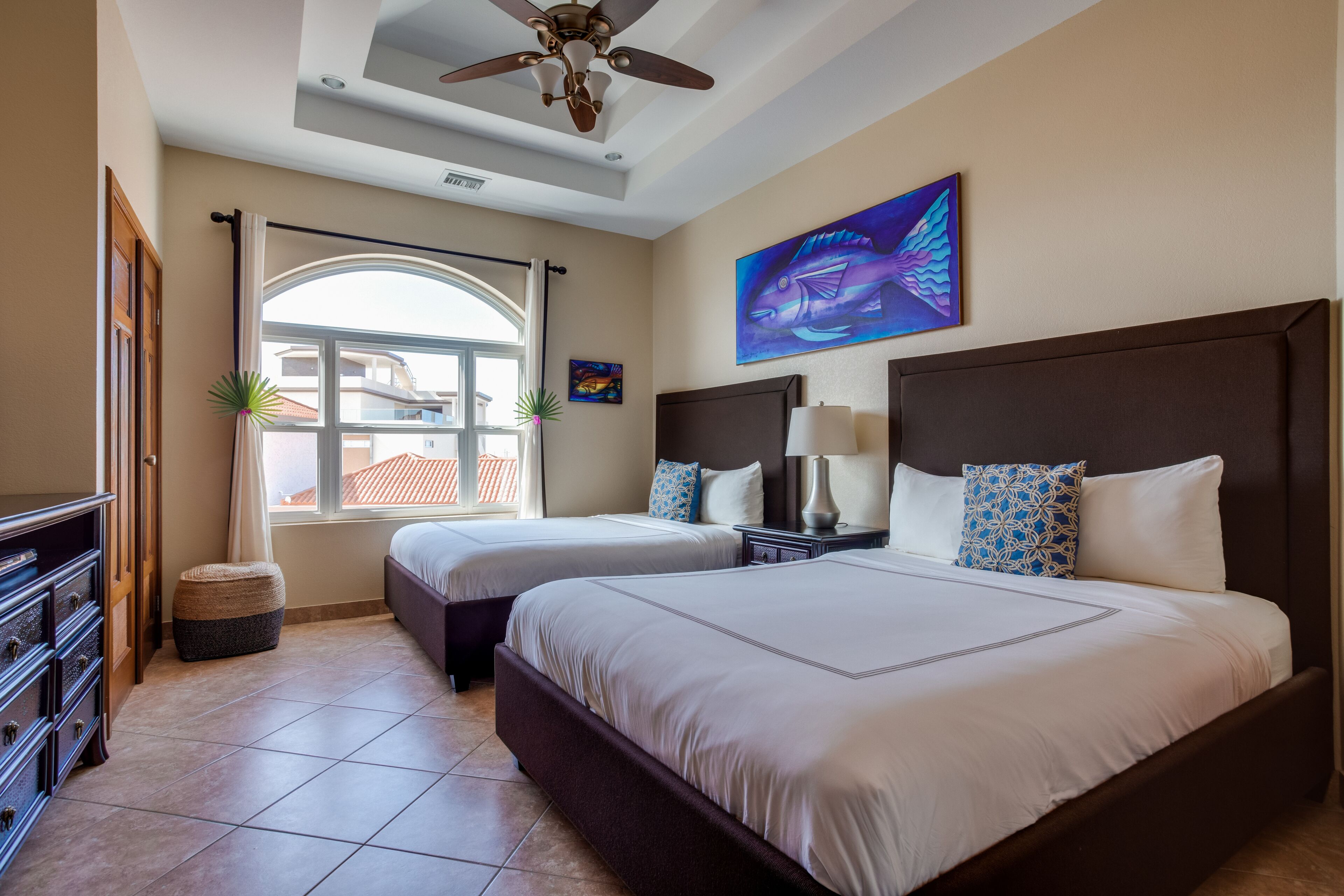 A hotel room with two beds, a ceiling fan, a large window with curtains, a painting of a fish, and a view of buildings outside.