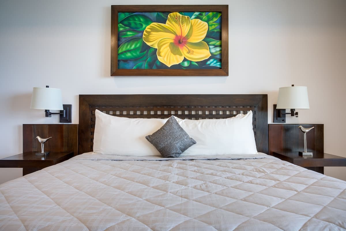 Una cama bien hecha con cubrecolcha acolchado, una sola almohada y una obra de arte floral enmarcada encima.