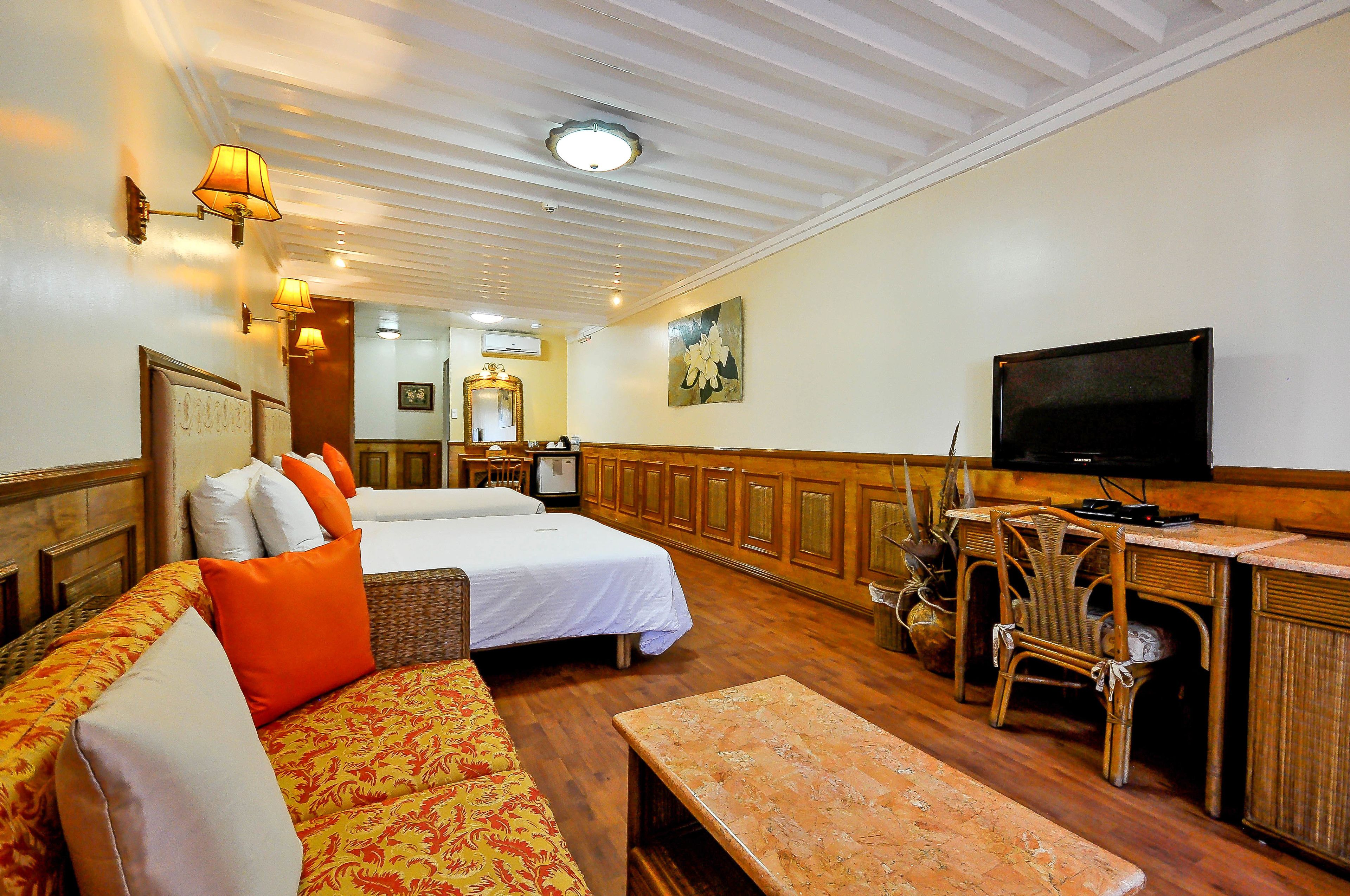 A hotel room with two beds, a sofa, a desk with a television, and a dining area with chairs.