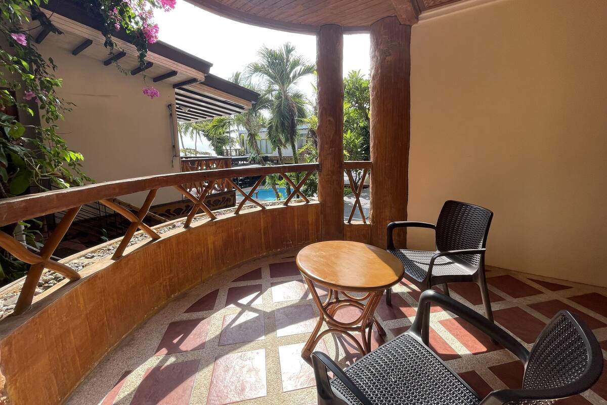 A covered outdoor seating area with a wooden table and chairs, a tiled floor, and a view of palm trees and a pool.