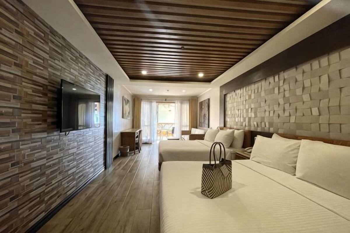 A hotel room with a wooden floor, a bed with white bedding, a TV mounted on the wall, and a brick accent wall.