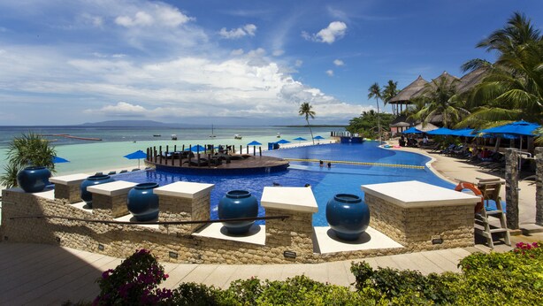A resort pool with a view of the ocean, surrounded by palm trees and lounge chairs. - The Bellevue Resort (Panglao)