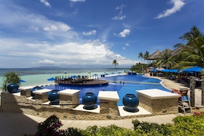 Piscina de resort con vista al océano, rodeada de palmeras y sillas de descanso.