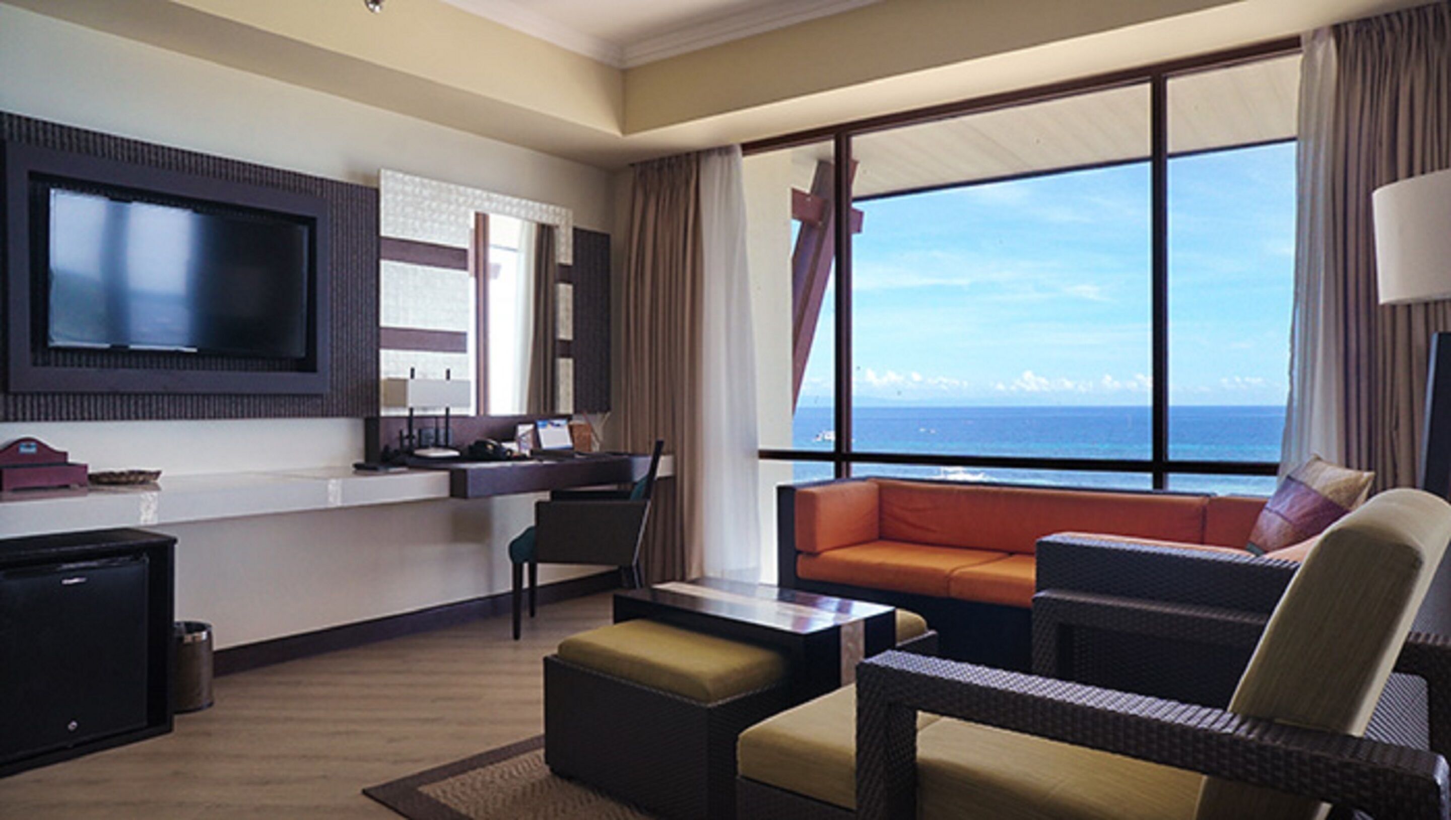 A hotel room with a large window offering a view of the ocean, a flat-screen TV mounted on the wall, a desk with a computer, and a seating area with a sofa and chairs.