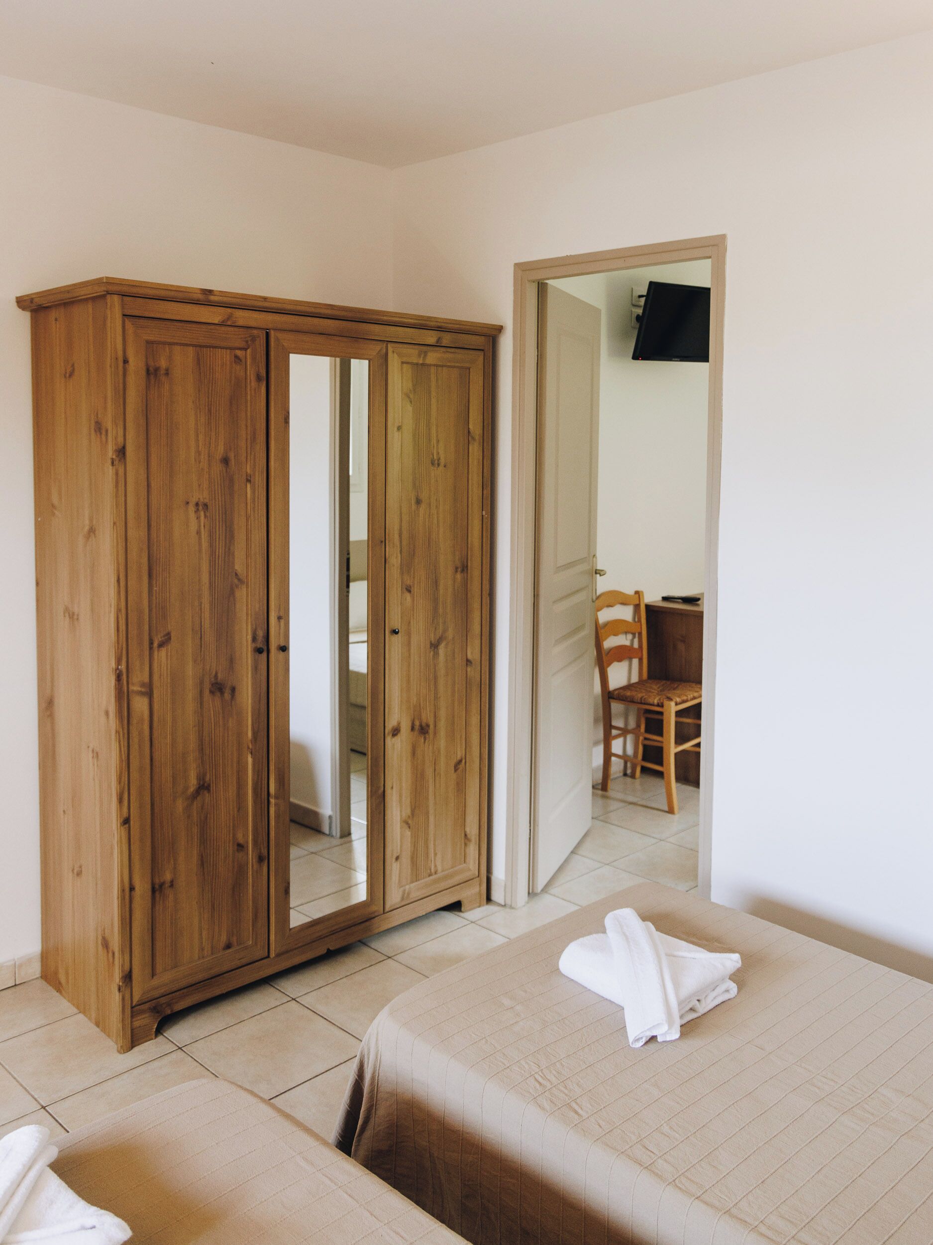 A bedroom with a wooden wardrobe, a bed with a beige bedspread, and a chair in the background.