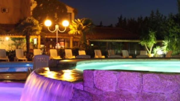A pool with a stone wall and a waterfall feature, surrounded by lounge chairs and a building with a warm exterior.