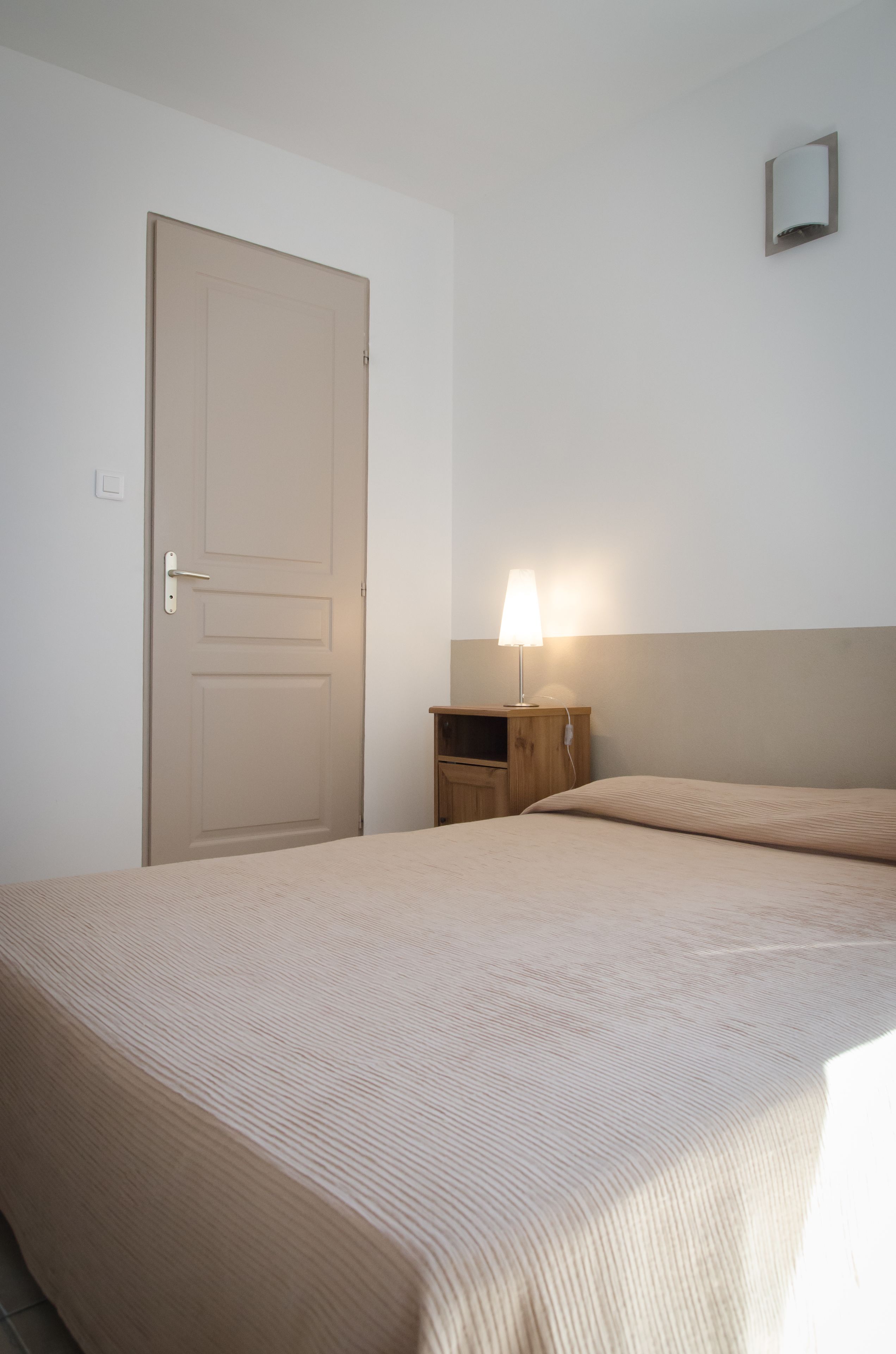 Un dormitorio con una cama, una mesita de noche con lámpara, una puerta y un pequeño aplique de pared.