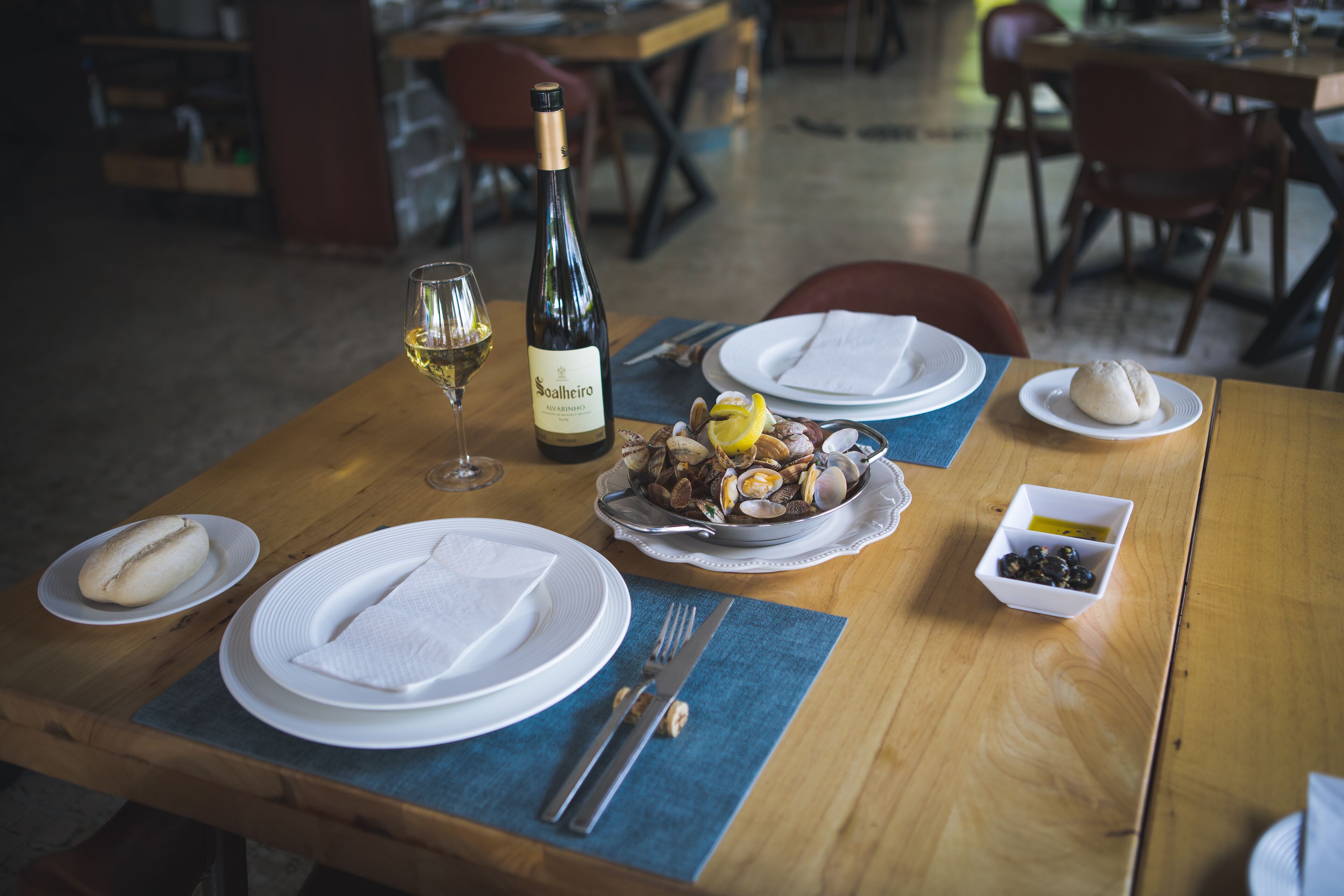 A dining table set with plates, cutlery, a bottle of wine, a bowl of clams, and a small dish of olives.