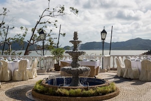 Un área de comedor al aire libre con una fuente central, mesas dispuestas para una comida y una vista panorámica de un lago al fondo.