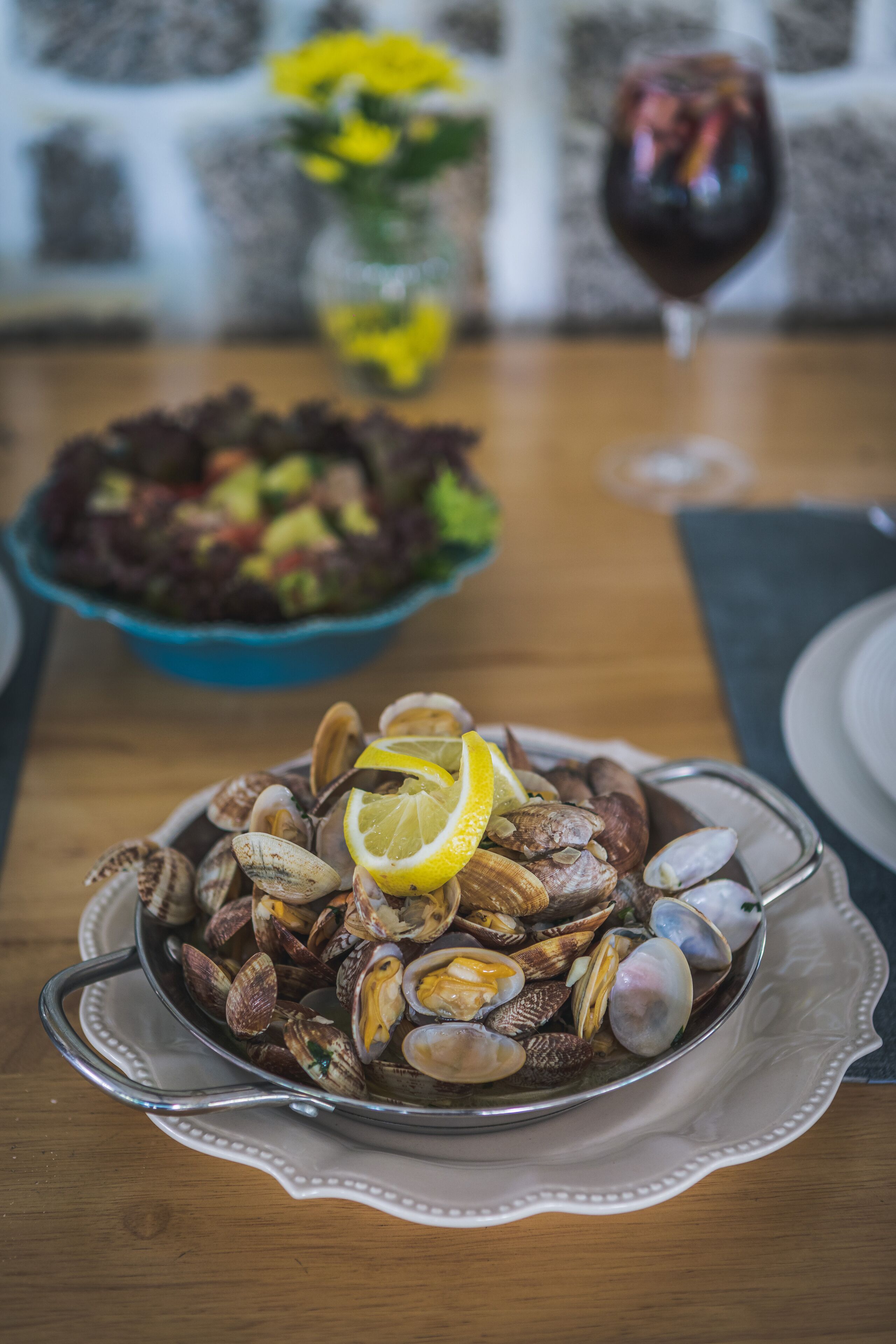 Una mesa de comedor con un bol de almejas adornado con rodajas de limón, una copa de vino tinto y un bol de ensalada al fondo.