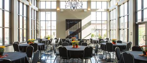 A spacious indoor area with multiple round tables and chairs, large windows, and a chandelier.