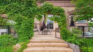 Una escalera de piedra conduce a un patio al aire libre con una mesa y sillas, rodeado de exuberante vegetación y un edificio al fondo.