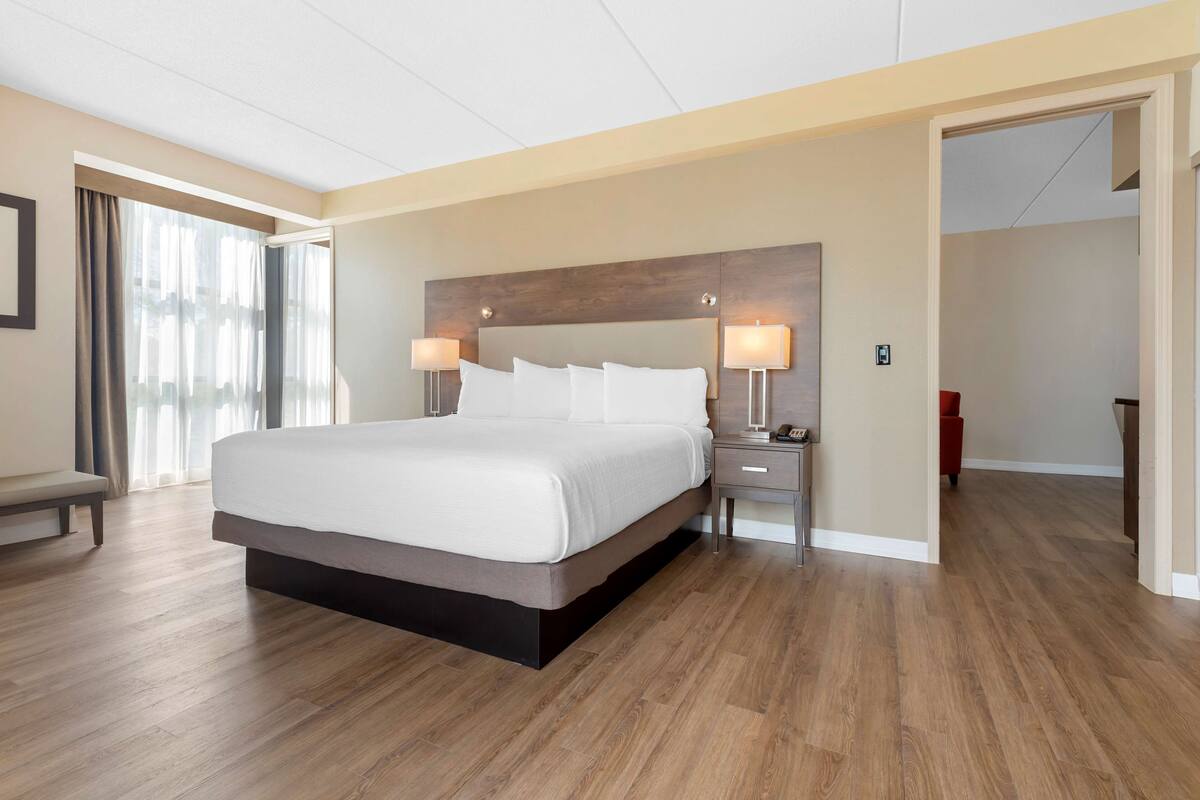 Una habitación de hotel moderna con una cama grande, lámparas de noche, un banco y vistas a través de una ventana con cortinas.