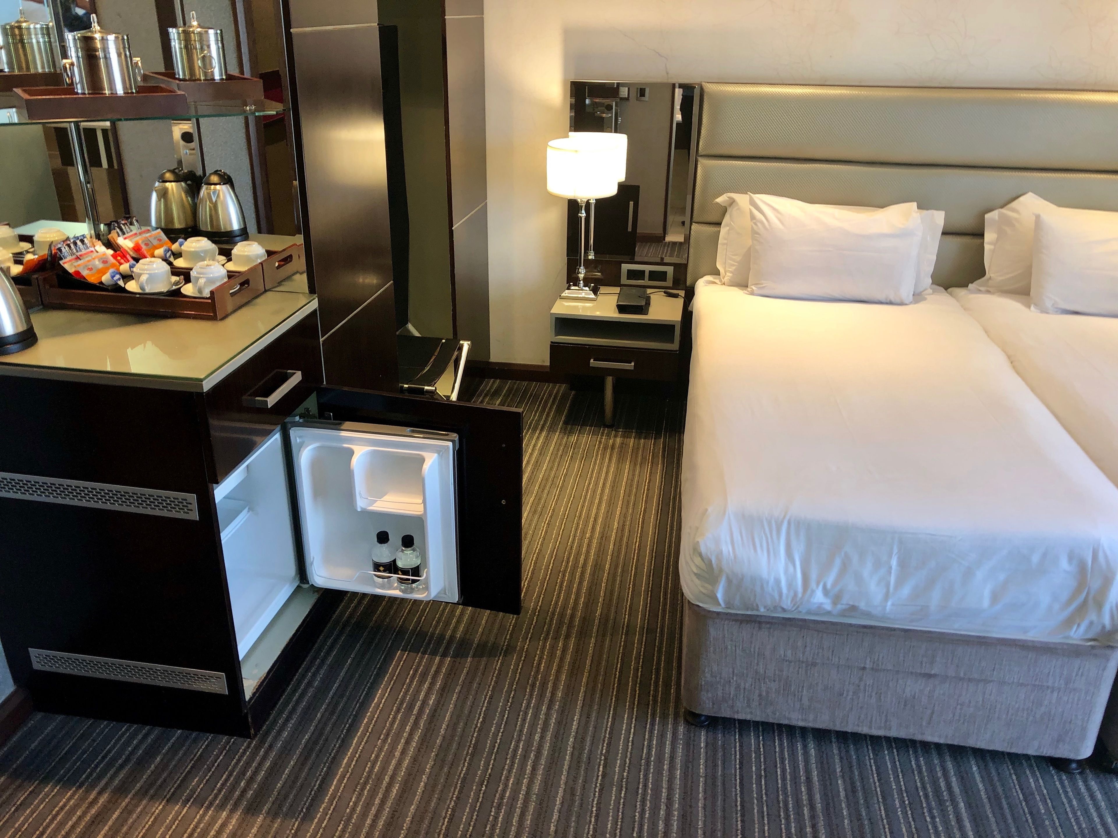 A hotel room with a bed, a desk with a coffee maker, a lamp, and a minibar.