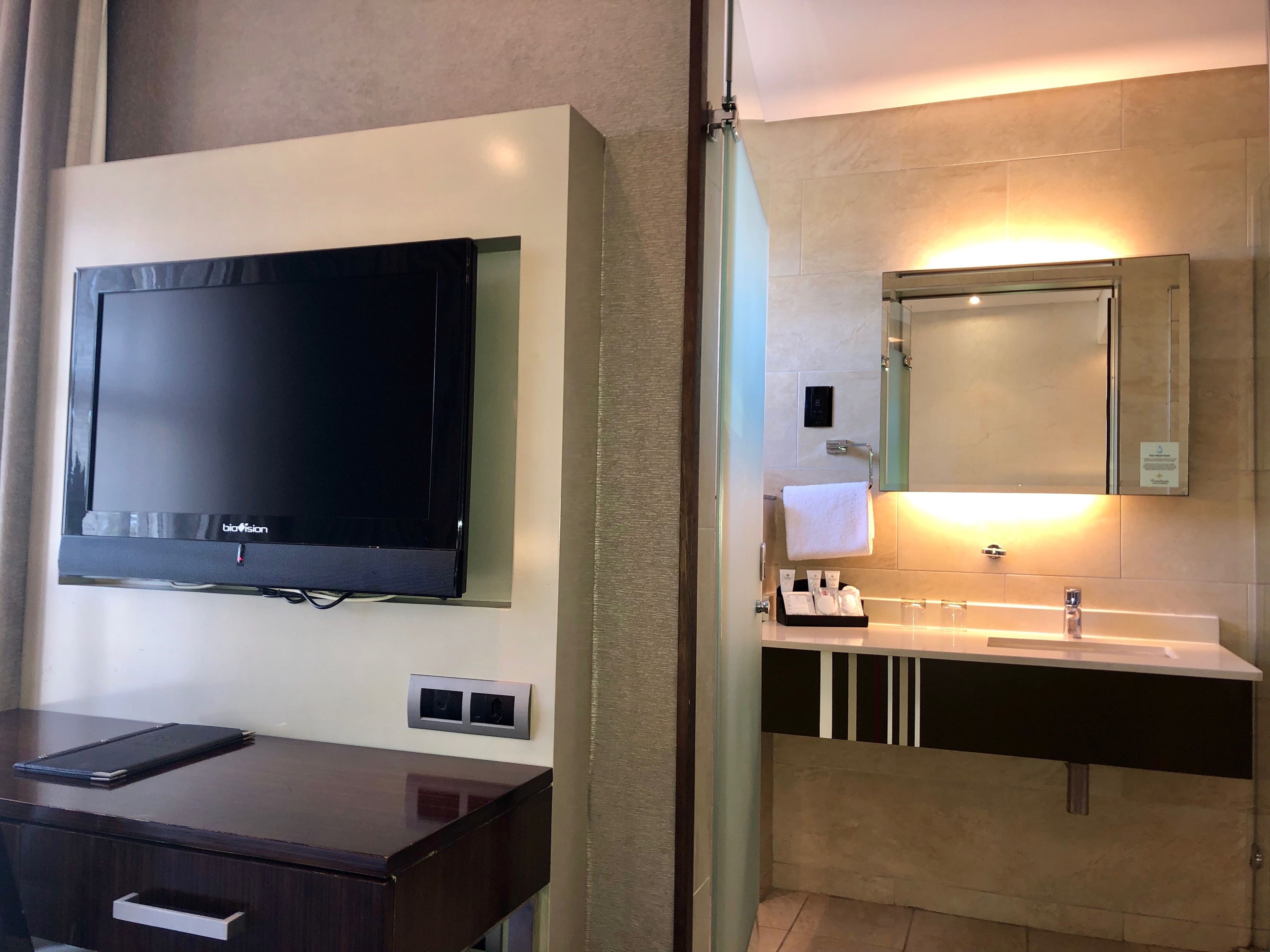 Un baño moderno con un televisor de pantalla plana montado en la pared, un tocador con lavamanos y una toalla colgada.