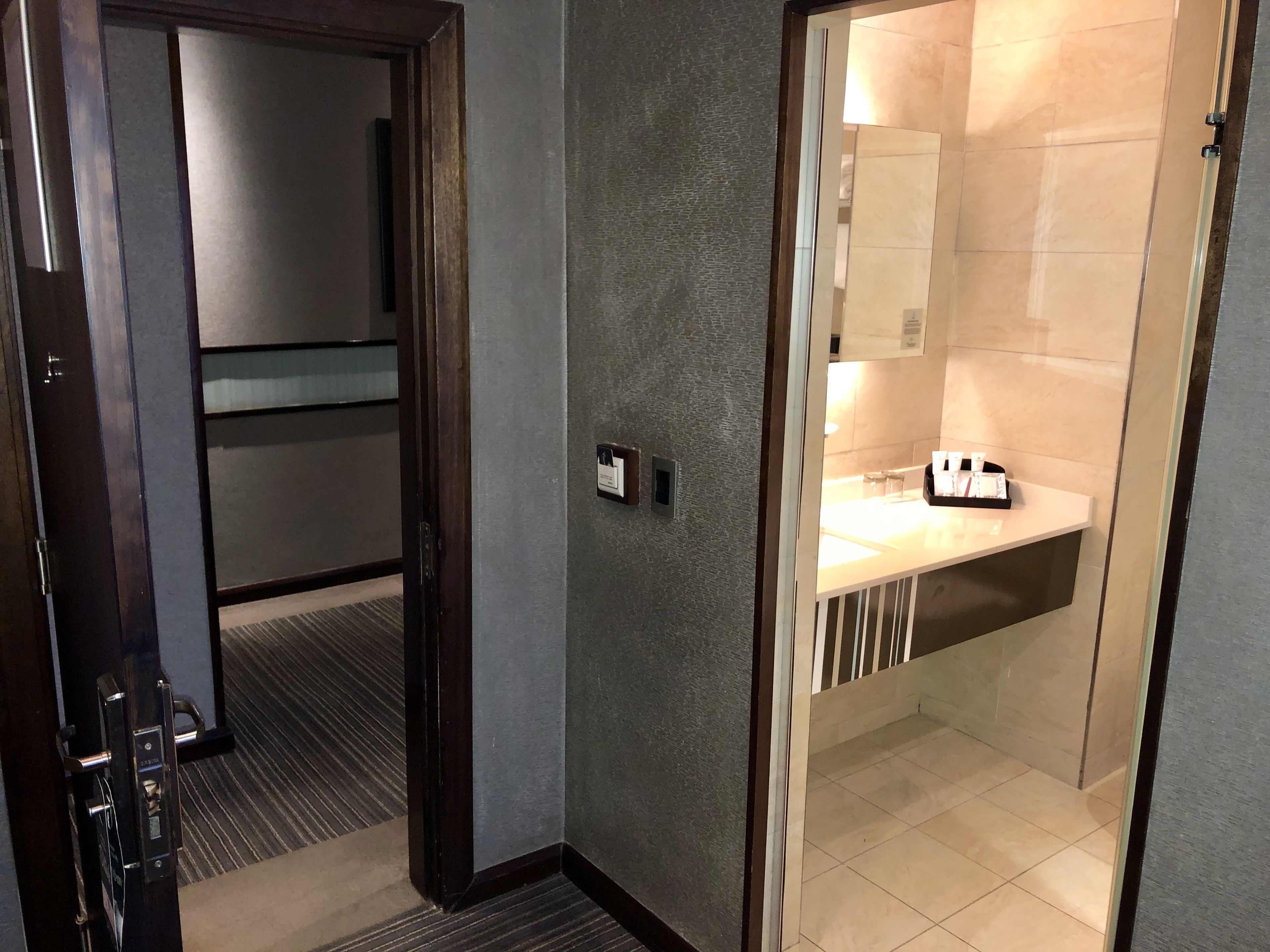 A hotel room with a modern design, featuring a dark wooden door, a textured grey wall, and a bathroom with a white sink and toiletries.