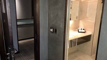 A hotel room with a modern design, featuring a dark wooden door, a textured grey wall, and a bathroom with a white sink and toiletries.