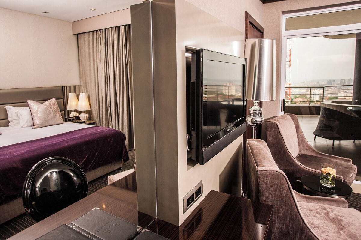 A hotel room with a bed, a TV, a seating area with a chair, and a view of the city.