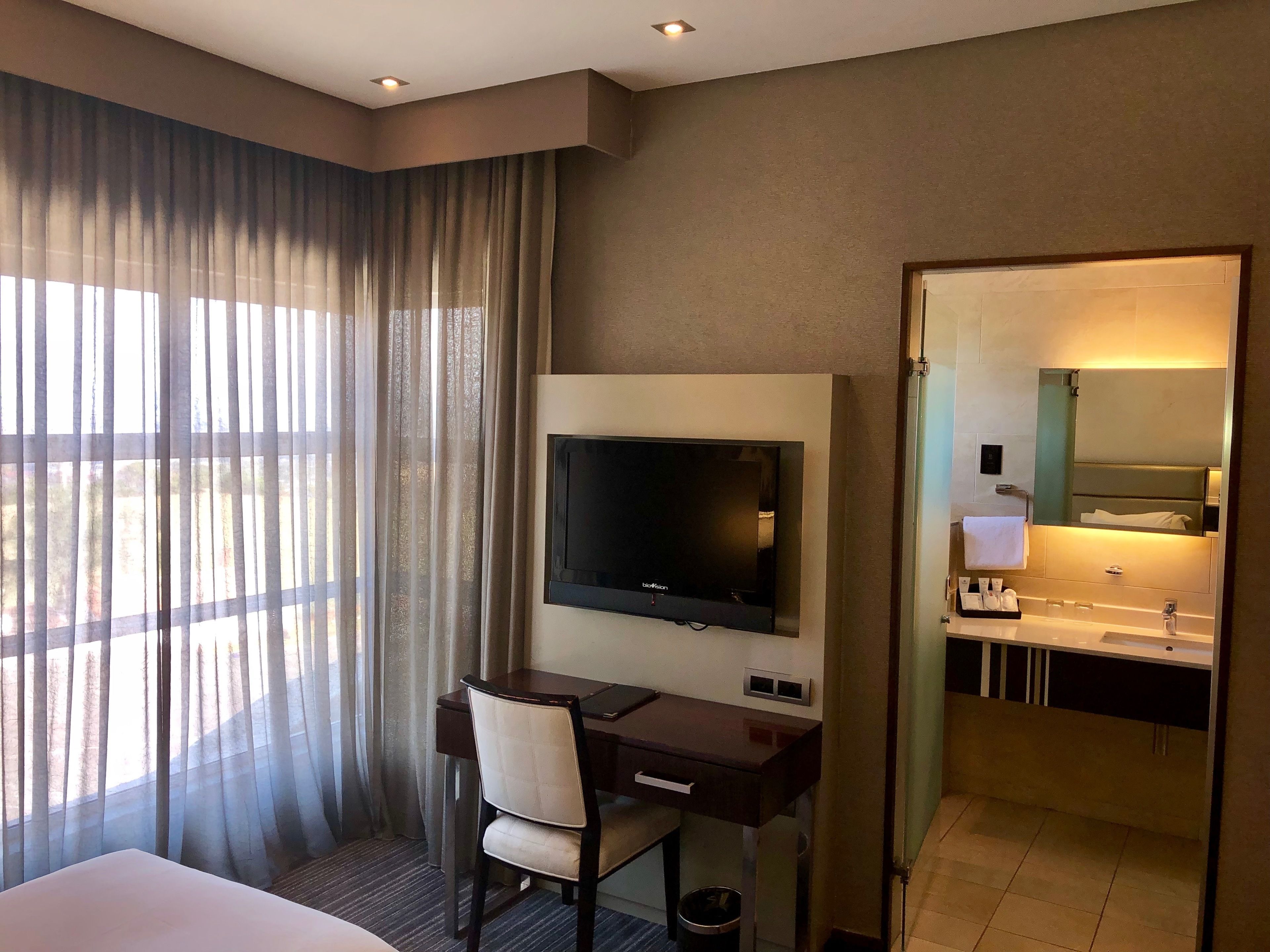 Ein Hotelzimmer mit einem Bett, einem Schreibtisch mit Stuhl, einem Fernseher, einem großen Spiegel und einem Badezimmer mit Waschbecken und Spiegel.