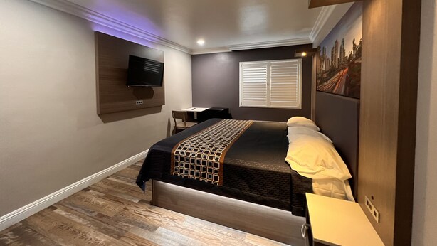 A modern hotel room with a bed, a desk, a chair, a TV, and a window with blinds. - Patio Motel (Gardena)