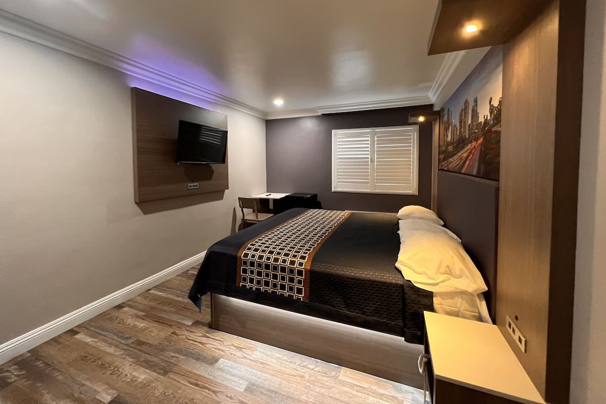 Quarto de hotel moderno com uma cama, uma escrivaninha, uma cadeira, uma televisão e uma janela com persianas.