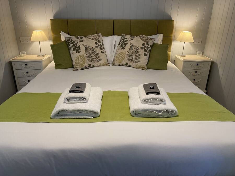Un lit bien fait, avec deux serviettes pliées et un téléphone de chaque côté, flanqué de tables de chevet avec des lampes et d’un plaid vert.