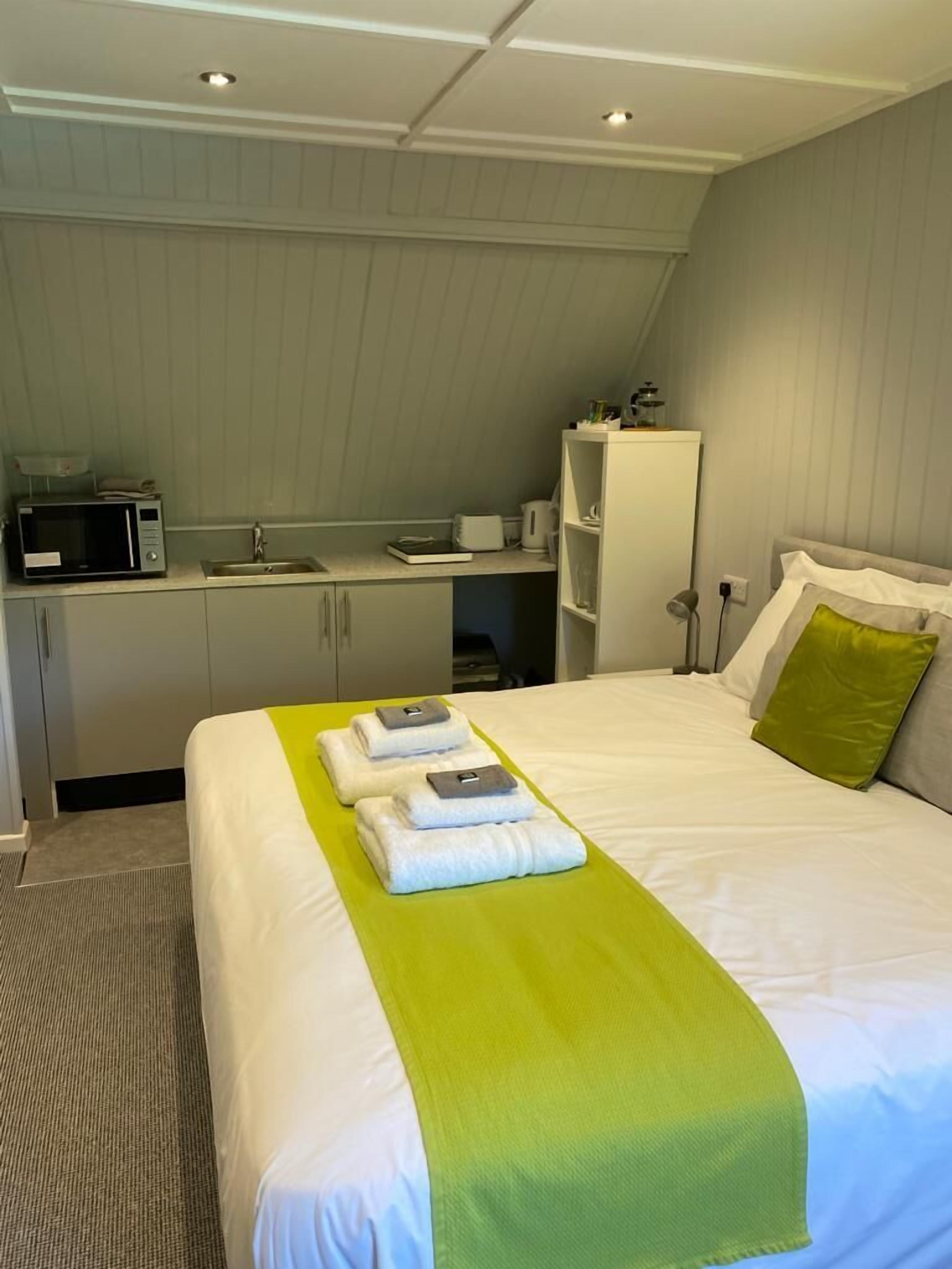 Una habitación de hotel con una cama bien hecha, una kitchenette pequeña y una mesita de noche con una lámpara.