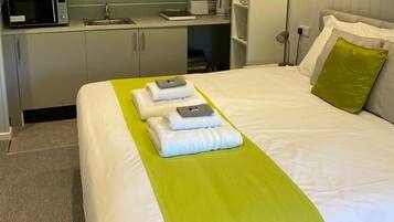 Una habitación de hotel con una cama bien hecha, una kitchenette pequeña y una mesita de noche con una lámpara.