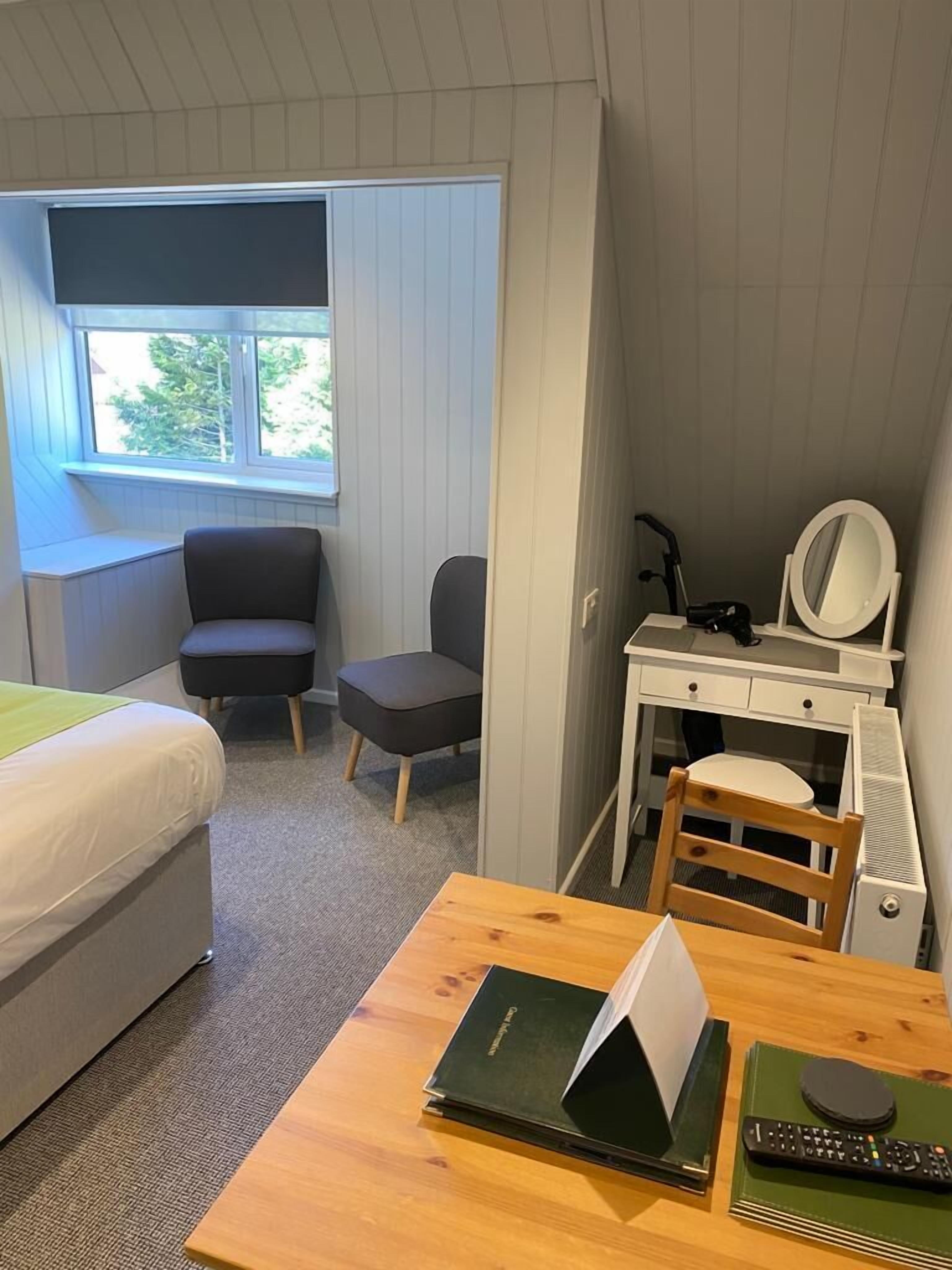 A hotel room with a bed, a desk with a chair, a small table with a book and remote control, and a window with a view of trees.