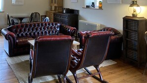 A spacious living room with exposed wooden beams, a leather sofa, matching armchairs, a dining table, and a radiator. - Riverside B & B (Leyburn)